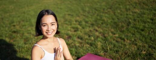 verticale tiro di giovane asiatico donna fare yoga, pratica consapevolezza, sorridente e guardare rilassato, seduta su gomma da cancellare stuoia nel parco foto