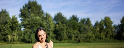 ritratto di asiatico ragazza dire Ciao su smartphone, onda a mobile Telefono mentre fare meditazione, in linea yoga classi su fresco aria nel parco foto