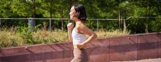 benessere e sport. asiatico fitness ragazza corridore, jogging nel parco, in esecuzione su strada nel ghette, sorridente felicemente foto