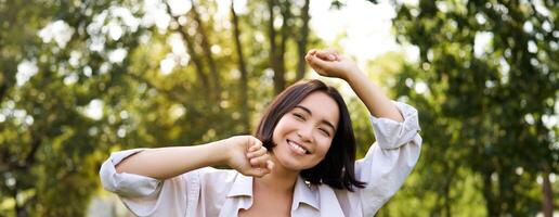 persone stile di vita. ritratto di giovane brunetta donna ballare, sorridente e ridendo, a piedi nel parco con mani sollevamento su alto, godendo estate giorno al di fuori foto