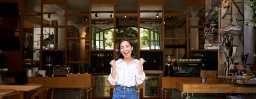 contento entusiasta asiatico ragazza, attività commerciale proprietario trionfante, apertura sua proprio ristorante, in piedi nel davanti di vuoto bar foto