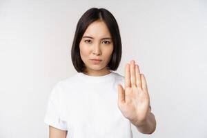 Immagine di asiatico ragazza mostrando fermare, vietare qualcosa, estendere uno braccio per mostrare vietando, tabù gesto, in piedi nel maglietta al di sopra di bianca sfondo foto