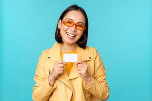 elegante giovane asiatico donna nel occhiali da sole, mostrando credito carta e sorridente, consigliare banca, senza contatto pagamento o sconti nel negozio, in piedi al di sopra di blu sfondo foto