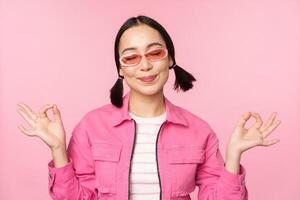 consapevolezza e benessere concetto. sorridente coreano ragazza nel elegante attrezzatura meditando, ascoltando mantra, detiene mani nel zen, pace posa, pratica yoga, in piedi al di sopra di rosa sfondo foto