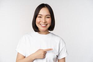 contento bellissimo asiatico ragazza mostrando annuncio pubblicitario su vuoto copia spazio, puntamento dito giusto e sorridente, dimostrando bandiera o azienda logo, bianca sfondo foto