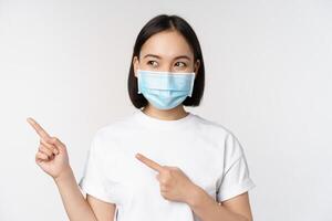giovane coreano donna nel medico viso maschera puntamento dita sinistra e guardare a logo, mostrando annuncio pubblicitario o striscione, in piedi al di sopra di bianca sfondo foto