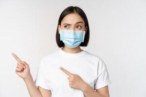 Immagine di asiatico donna nel medico maschera a partire dal covid, guardare confuso sinistra, puntamento a logo, mostrando annuncio, in piedi al di sopra di bianca sfondo foto