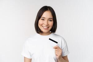 ritratto di coreano sorridente ragazza, banca cliente, mostrando credito carta con contento viso, in piedi contro bianca sfondo foto