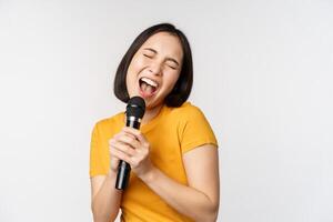 contento asiatico ragazza danza e cantando karaoke, Tenere microfono nel mano, avendo divertimento, in piedi al di sopra di bianca sfondo foto