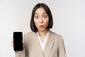Immagine di asiatico aziendale donna mostrando App interfaccia, mobile Telefono schermo, fabbricazione sorpreso viso espressione, oh, in piedi al di sopra di bianca sfondo foto