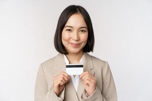 sorridente ufficio impiegato, asiatico aziendale donna mostrando credito carta, in piedi al di sopra di bianca sfondo nel beige completo da uomo foto