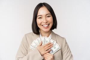 contento asiatico donna d'affari Tenere Contanti, abbracciare dollari i soldi e sorridente, in piedi al di sopra di bianca sfondo nel completo da uomo foto
