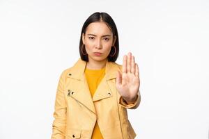 grave e ha riguardato asiatico ragazza estendendosi palma mano, fermare gesto, vietare qualcosa, proibire, in piedi al di sopra di bianca studio sfondo foto