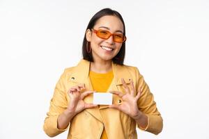 elegante attraente asiatico ragazza nel occhiali da sole, mostrando credito carta e sorridente, in piedi contento contro bianca studio sfondo foto