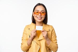 ritratto di sorridente coreano femmina modello nel occhiali da sole, mostrando credito carta, in piedi al di sopra di bianca sfondo foto