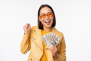 microcredito e i soldi concetto. elegante asiatico giovane donna nel occhiali da sole, ridendo felice, Tenere dollari Contanti, in piedi al di sopra di bianca sfondo foto
