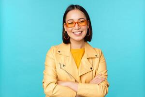 elegante coreano donna nel occhiali da sole e giallo giacca, attraversare braccia, sorridente e guardare fiducioso a telecamera, in piedi contro blu sfondo foto