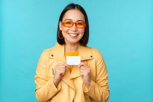 giovane bellissimo asiatico donna mostrando credito carta, sorridente, la scelta banca, in piedi al di sopra di blu sfondo foto