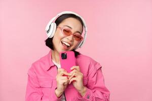 elegante asiatico ragazza nel cuffia, ascoltando musica e assunzione fotografie su mobile Telefono, utilizzando smartphone, in piedi al di sopra di rosa sfondo