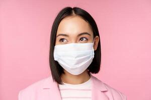 vicino su ritratto di giapponese aziendale donna nel medico viso maschera a partire dal covid19, guardare sinistra a logo, vendita promo, in piedi al di sopra di rosa sfondo foto