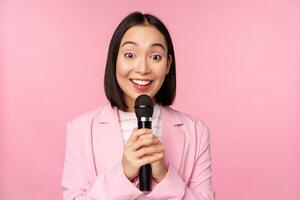 asiatico donna d'affari dando discorso, Tenere microfono e sorridente, in piedi nel completo da uomo al di sopra di rosa sfondo foto