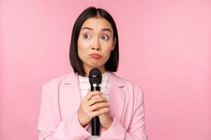 nervoso femmina imprenditore dando discorso con microfono, Tenere mic e guardare a parte con ansia, in posa nel completo da uomo al di sopra di rosa sfondo foto