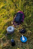 escursionismo, all'aperto attività. campeggio cucina utensili menzogna nel il erba un' termos, un' tazza, un' stufa, un' ciotola e un' zaino. turista prima colazione. cucinando nel natura. foto