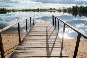 panorama di il molo di un' silenzioso lago su un' soleggiato giorno. un' di legno molo a il acqua superficie, nuvole siamo riflessa nel il acqua, e un' foresta cresce intorno a. foto