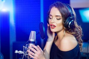 ritratto di attraente donna con Perfetto trucco registrazione un' canzone nel un' professionale studio. bella femmina con cuffie e microfono cantando un' canzone. foto