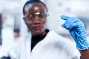 campione bicchiere diapositiva nel messa a fuoco mentre chimico è guardare a esso durante laboratorio indagine. africano assistenza sanitaria scienziato nel biochimica laboratorio indossare sterile attrezzatura. foto