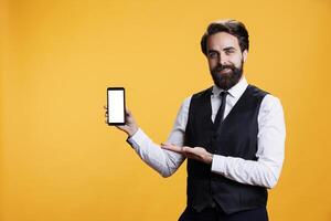 maschio Cameriere Spettacoli bianca schermo nel studio, presentazione isolato copyspace modello su smartphone Schermo. professionale elegante maggiordomo Tenere vuoto modello schermo al di sopra di giallo sfondo. foto