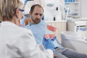 specialista stomatologo medico con viso maschera Tenere mascella ceramica modello spiegando orale igiene per uomo paziente durante odontoiatria visita medica nel stomatologiche ufficio camera. stomatologia concetto foto