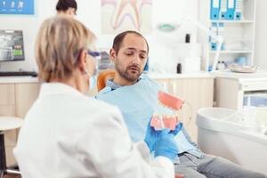 stomatologo infermiera spiegando per malato uomo il chirurgia utilizzando gesso modello di il dentista dentista Tenere modello di scheletro di denti, campione di umano mascella parlando. durante durante stomatologia ospedale foto