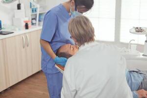 malato uomo seduta su dentale sedia mentre anziano medico assunzione cura di dente Salute. stomatologo A proposito di per paziente mentre infermiera pulizia bocca durante dentale visita medica nel stomatologia clinica foto