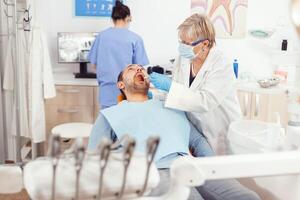 ortodontista anziano medico fare mal di denti trattamento su malato paziente preparazione chirurgia Strumenti. uomo dire bugie su ortodontico sedia nel ospedale stomatologiche ufficio durante medico procedura foto