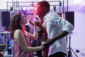 romantico giovane coppia Tenere mani e danza insieme mentre discoteca. diverso fidanzato e fidanzata partying e in movimento su pista da ballo nel discoteca illuminato con luci foto