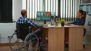 squadra di videografi Lavorando a video progetto la creazione di contenuto, uomo blogger seduta nel sedia a rotelle nel moderno azienda ufficio. Disabilitato libero professionista la modifica metraggio post produzione nel studio. foto