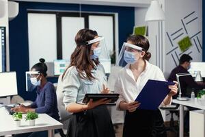 donne d'affari con protezione maschere contro coronavirus in piedi nel posto di lavoro parlando di azienda finanziario dati Tenere digitale tavoletta. multietnico attività commerciale squadra Lavorando rispetto sociale distanza durante globale pandemia con coronavirus. foto