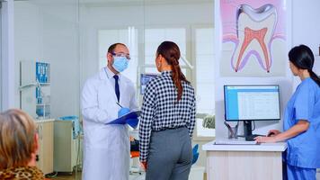 dentista medico interrogando donna e assunzione Appunti su appunti in piedi nel in attesa la zona. giovane paziente spiegando dentale problema per stomatologo A proposito di nel affollato ricezione di clinica. foto