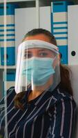 manager donna con visiera e protezione maschera guardare a telecamera sorridente nel nuovo normale attività commerciale ufficio. libero professionista Lavorando nel finanziario azienda rispetto sociale distanza durante globale pandemia. foto