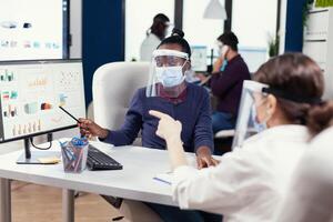 africano manager spiegando progetto grafico per dipendente indossare viso maschera. multietnico squadra Lavorando nel azienda con nuovo normale rispetto sociale distanza perché di globale pandemia con coronavirus. foto