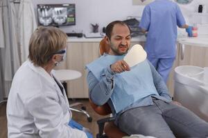 paziente Tenere specchio guardare a denti procedura dopo dentale chirurgia durante stomatologia visita medica nel stomatologiche ufficio camera. dentista anziano donna spiegando orale igiene. concetto di medicina foto