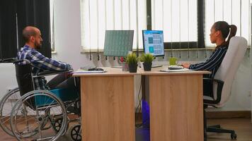 donna d'affari analizzando finanziario statistica parlando con portatori di handicap collaboratore seduta nel sedia a rotelle controllo grafici su scrivania nel edificio ufficio. Disabilitato uomo d'affari utilizzando moderno tecnologia foto