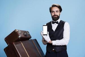 elegante fattorino usi mobile Telefono con bianca Schermo, Tenere dispositivo nel davanti di telecamera e indossare elegante concierge Abiti. professionale portiere Spettacoli vuoto copyspace schermo. foto