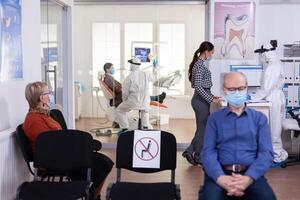 dentista infermiera vestito nel ppe completo da uomo con viso shiled discutere con paziente nel stomatologia in attesa camera. persone conservazione sociale distanziamento come prevenzione durante coronavirus scoppio. foto