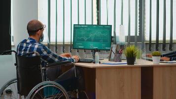 Disabilitato uomo d'affari seduta nel sedia a rotelle con protezione maschera pulizia mani prima controllo finanziario dati nel attività commerciale moderno ufficio. portatori di handicap libero professionista con visiera rispetto sociale distanza foto