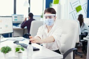 imprenditore indossare viso maschera e viso scudo utilizzando gel per mano hygine durante globale pandemia con covid19. donna d'affari nel nuovo normale posto di lavoro disinfezione mentre colleghi Lavorando nel sfondo. foto