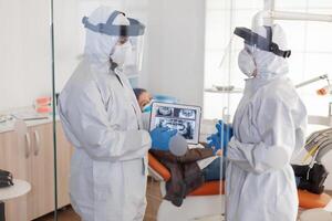 squadra di medici indossare pieno virus protezione uniforme in piedi vicino paziente, analizzando denti digitale raggi X utilizzando tavoletta pianificazione trattamento. concetto di nuovo normale dentista visitare nel coronavirus scoppio. foto