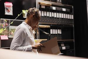 africano americano donna avvocato studiando crimine importa file nel investigatore ufficio. legge rinforzo professionale analizzando legale competenza rapporto nel investigatore camera a notte tempo foto