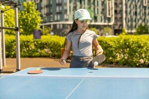 giovane adolescente ragazza giocando ping pong. lei detiene un' palla e un' racchetta nel sua mani. giocando tavolo tennis all'aperto nel il cortile foto